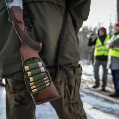 Myśliwy w lesie, z bronią i amunicją w pasie. W tle inni uczestnicy polowania w kamizelkach odblaskowych.