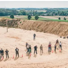 Grupa ludzi na strzelnicy z piaszczystym podłożem i nasypem ziemnym w tle.
