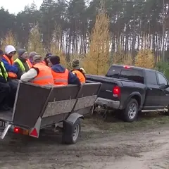 Grupa osób w kamizelkach odblaskowych jedzie przyczepą ciągniętą przez pickupa w lesie.