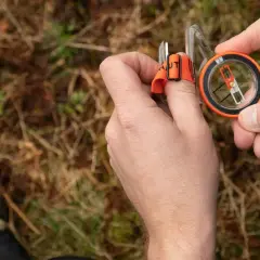Dłonie trzymające kompas na tle suchej trawy i butów trekkingowych.