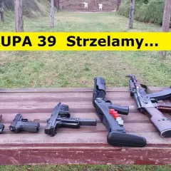 Na drewnianym stole leżą różne rodzaje broni palnej, w tle strzelnica.