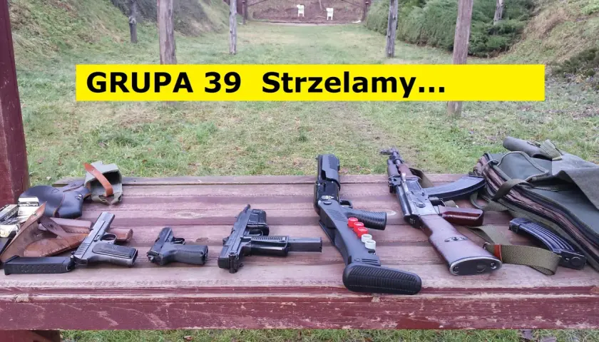 Na drewnianym stole leżą różne rodzaje broni palnej, w tle strzelnica.