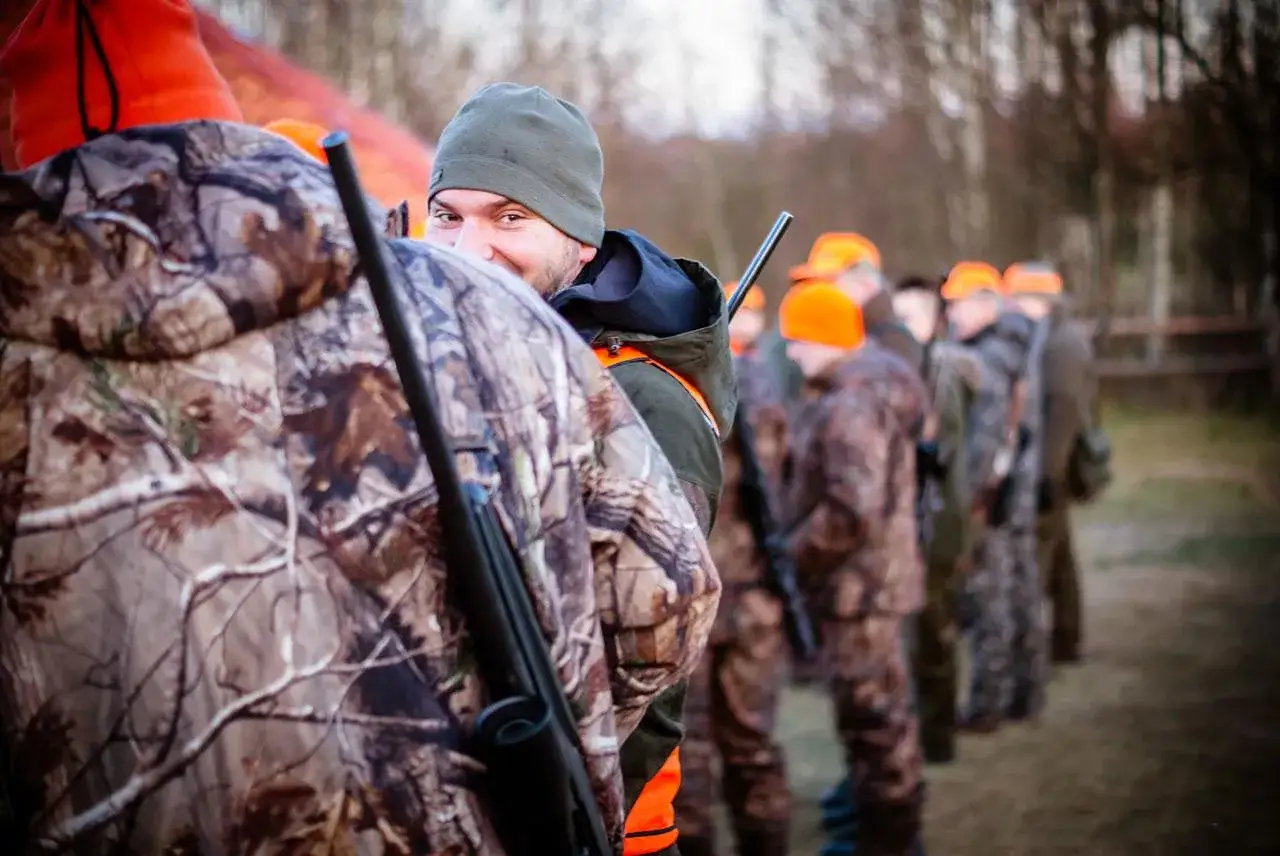 Myśliwi w kamuflażu i pomarańczowych czapkach, z bronią, czekają na sygnał. Tak powinien przemieszczać się myśliwy, zachowując ostrożność.